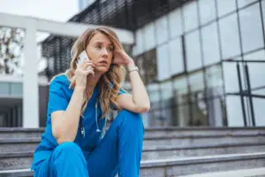 Caregiver in scrubs sitting on steps, looking stressed while holding a phone, highlighting caregiver burnout and mental health challenges.