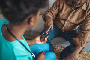 A home nurse helps an older man check his blood sugar using a glucose monitor during an in-home care visit.